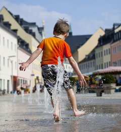 Foto, das einen kleinen Jungen zeigt, der in einer Stadt durch einen Springbrunnen läuft.